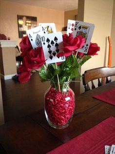 a vase filled with red flowers on top of a wooden table next to playing cards