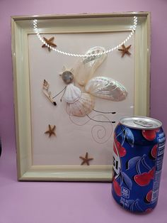 a beer can sitting next to a framed artwork with shells and starfish on it