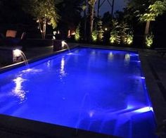 an outdoor swimming pool lit up at night with lights on the side and trees in the background