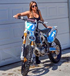 a woman standing next to a dirt bike in front of a garage door with her hands on the handle bars