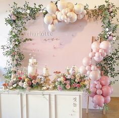 a table topped with lots of pink and white balloons next to a wall covered in greenery