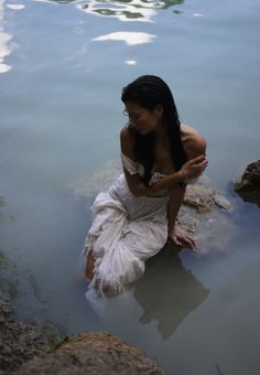 a woman in a white dress sitting on rocks in the water with her hands together