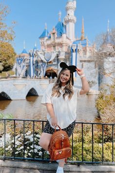 a woman posing in front of a castle with her hand on her hat and purse