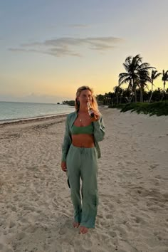 a woman standing on top of a sandy beach next to the ocean holding a drink