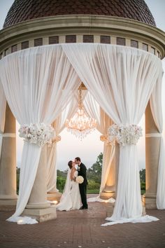 a bride and groom standing under an outdoor gazebo with white drapes on it