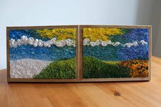 two pieces of art are sitting on a wooden table, one is painted with grass and the other has clouds