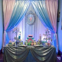 a table topped with lots of cakes and desserts next to a window covered in blue drapes