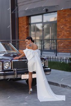 a woman standing next to a car in front of a building wearing a white dress