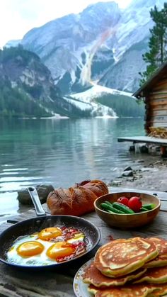 pancakes, eggs and vegetables sit on a picnic table overlooking a lake with mountains in the background