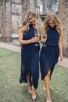 two women in blue dresses holding wine glasses and looking at each other while standing on the grass