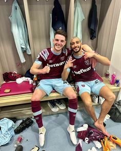 two soccer players sitting on a bench in the dressing room with their arms around each other