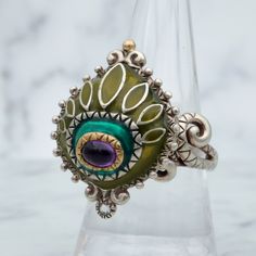 This impressive ring by Barbara Bixby is made of sterling silver with 18k gold accents and features shades of green enamel with an amethyst eye, quartz drop and highly detailed engravings on the band and undercarriage. This ring is finished with stunning swirled "octopus" arms on the shoulders. It is a size 9, weighs 14.3 grams and the face measures 29x22mm. It is in great vintage condition with light wear and is marked as shown.  *Matching earrings available Green Enamel Ring For Anniversary, Unique Green Enamel Ring For Anniversary, Unique Green Enamel Anniversary Ring, Unique Enamel Rings For Anniversary, Unique Collectible Enamel Ring, Unique Enamel Rings With Gemstone, Unique Enamel Sterling Silver Ring For Anniversary, Unique Enamel Ring In Sterling Silver For Anniversary, Unique Enamel Gemstone Ring