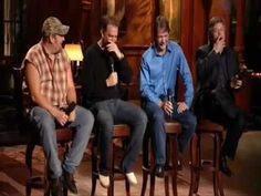 four men sitting on stools in front of a painting and one holding a microphone