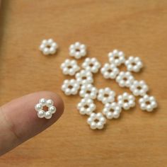 a hand holding a tiny white beaded flower on top of a wooden table next to some beads