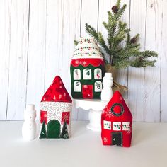 three red and green christmas houses sitting on top of a white table next to a pine tree