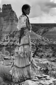 a woman standing on top of a rock covered hillside next to a desert area with mountains in the background