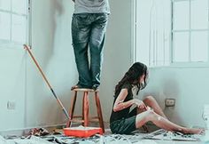 a man and woman standing on top of a ladder in a room with paint all over the floor