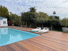 a wooden deck next to a swimming pool with lounge chairs on the side and trees in the background