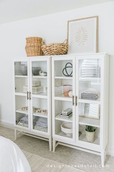 a white bookcase with glass doors and baskets on top is in the corner of a bedroom