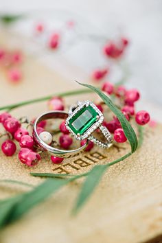 an emerald ring sitting on top of a piece of wood with pink flowers in the background