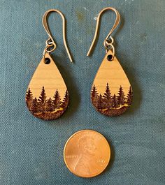 two wooden earrings with trees on them next to a quarter penny and blue fabric background