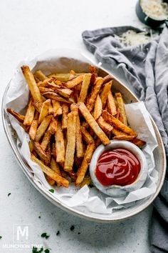 french fries with ketchup in a white bowl on a gray napkin next to a cup of ketchup