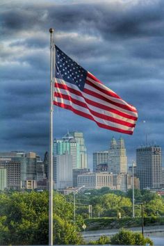 an american flag flying in front of a city skyline with dark clouds and blue sky