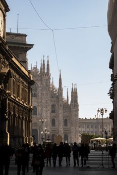 many people are walking down the street in front of an old building with tall spires