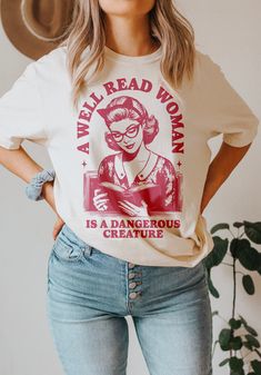 Celebrate the power of knowledge and embrace the spirit of rebellion with this shirt design that boasts a vintage 1950s illustration of a woman immersed in a book. As she captivates the era's charm, the surrounding text boldly declares, "A well-read woman is a dangerous creature." Ideal for book lovers, feminists, and banned book activists, this design encapsulates the strength and influence that come with intellectual prowess.  The juxtaposition of the retro aesthetic with the empowering messag Bookish Crew Neck Shirt With Graphic Print, Relaxed Fit Graphic Print Shirt In Bookish Style, Relaxed Fit Graphic Print Shirt With Bookish Style, Cotton Slogan T-shirt, Bookish Cotton Shirt With Graphic Print, Bookish Cotton Tops With Character Print, Literary Cotton Relaxed Fit T-shirt, Literary Style Cotton T-shirt With Relaxed Fit, Literary Graphic Print Crew Neck T-shirt