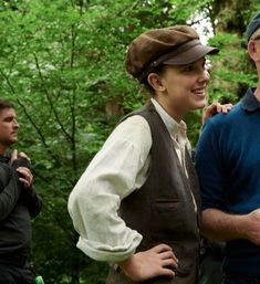 two men standing next to each other in front of some trees and one man is wearing a hat