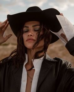 a woman with long hair wearing a black hat and holding her hands up to her head