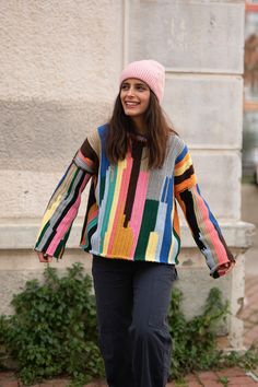 a woman is walking down the sidewalk wearing a colorful sweater and pink knitted hat