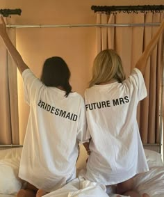 two women in matching white shirts sitting on a bed with their arms behind the head