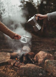 two people holding coffee cups over a campfire