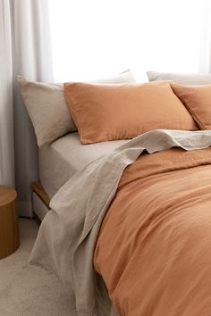 a bed with orange sheets and pillows in front of a white curtain, next to a small wooden side table