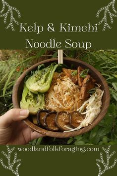 a hand holding a wooden bowl filled with noodles and vegetables, text reads keep & kimchii noodle soup