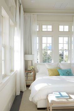 a white bed sitting in a bedroom next to two windows with lots of light on them