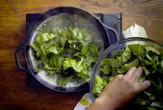 two bowls filled with lettuce on top of a wooden table next to a person's hand