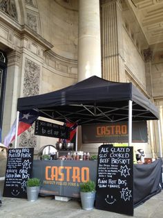 an outdoor food stand with chalk writing on the front and side walls, along with potted plants