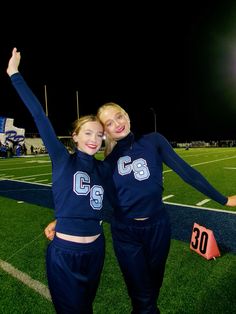 two cheerleaders pose for a photo on the football field at night with their arms in the air