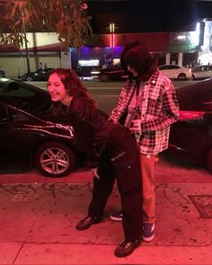 two people standing next to each other in front of a parked car on a city street