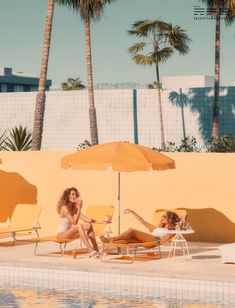 two women lounging on lounge chairs next to a swimming pool with palm trees in the background