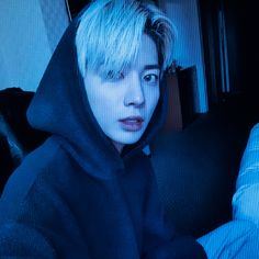 a person with blonde hair wearing a black hoodie and looking at the camera while sitting on a couch