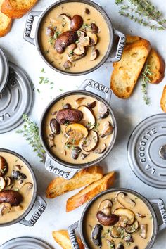 four pans filled with mushroom soup on top of a white table next to bread