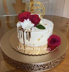 a white cake with red roses on top and gold decoration is sitting on a table