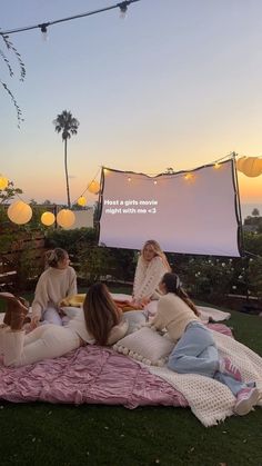 four women sitting on a blanket in front of a movie screen at sunset with paper lanterns