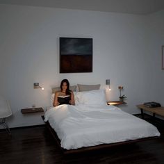 a woman sitting on top of a bed reading a book in a room with white walls