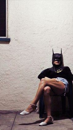 a woman dressed as batman sitting on a chair in front of a building with her legs crossed