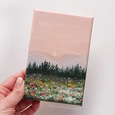 a hand holding up a small painting with trees in the background and flowers on the ground