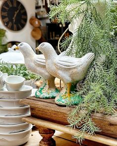 two white birds sitting on top of a wooden table next to plates and cups in front of a clock